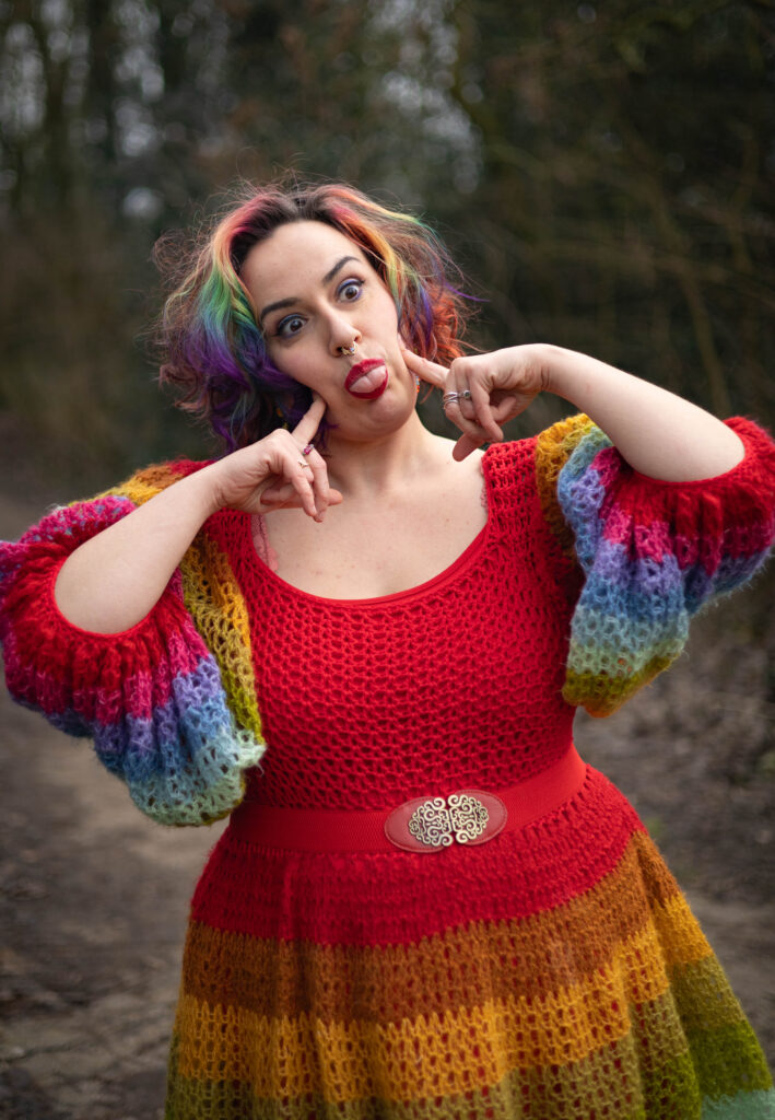 Jopie is een wit non-binair persoon met halflang haar in regenboogkleuren met een matchende gehaakte jurk aan, en trekt een gek gezicht met diens tong uitgestoken.